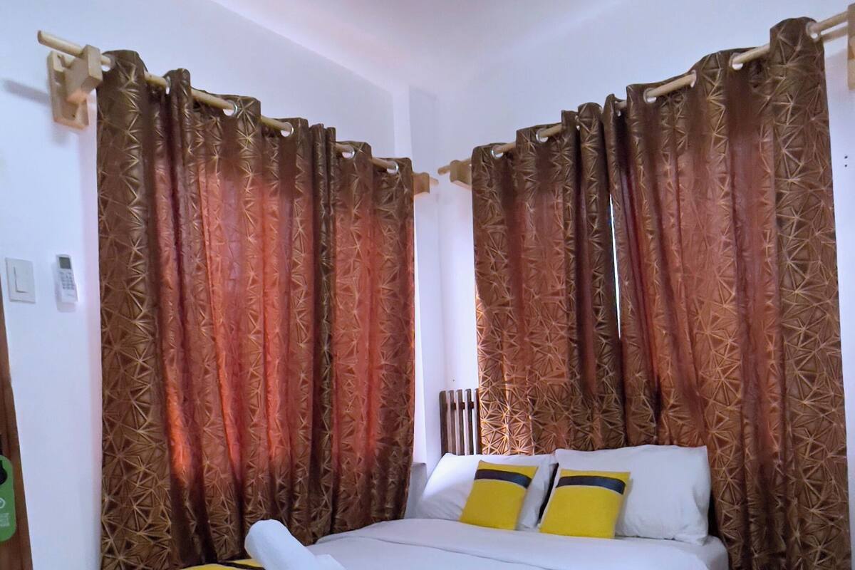 Una cama bien hecha con una colcha a rayas amarilla y azul, cortinas estampadas marrones y un cojín blanco.