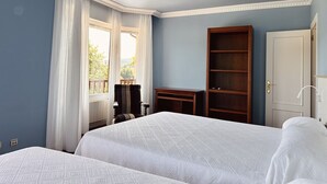 A hotel room with two beds, a wooden wardrobe, a desk, and a view of greenery through a window with white curtains. - El Rancho - 13801 (Güemes)