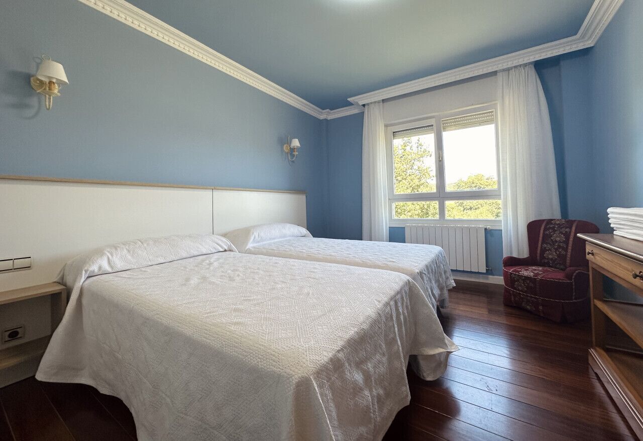 Een kamer met twee bedden, een raam met witte gordijnen, een stoel en een dressoir.