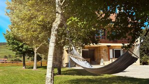 A hammock hanging between two trees in a grassy area with a building in the background. - El Rancho - 13801 (Güemes)