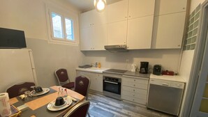 Cocina compacta con alacenas blancas, horno integrado, lavavajillas y cafetera. Hay una mesa de comedor con sillas y una ventana encima.