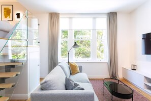 A modern living room with a grey sofa, a glass staircase, a large window with curtains, a floor lamp, and a flat-screen TV. - Coleherne Road Residence VI (London)