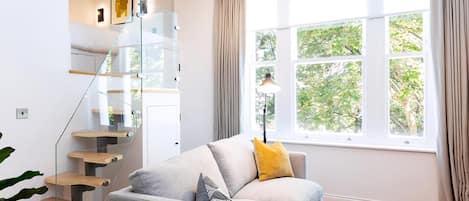 A modern living room with a grey sofa, a glass staircase, and a large window with curtains.