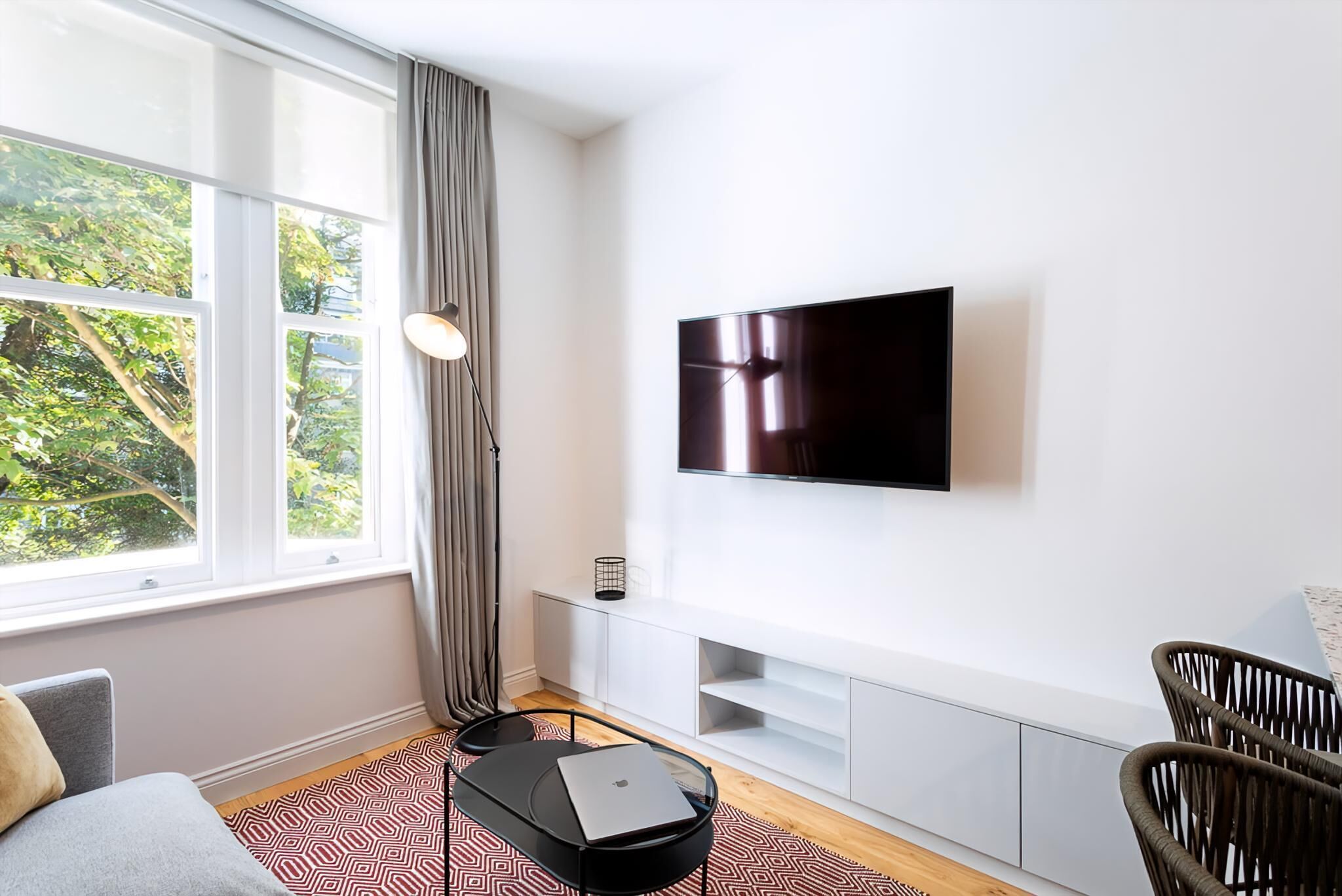 A modern living room with a large window, a flat-screen TV mounted on the wall, a sofa, a coffee table with a laptop, and a wicker chair.