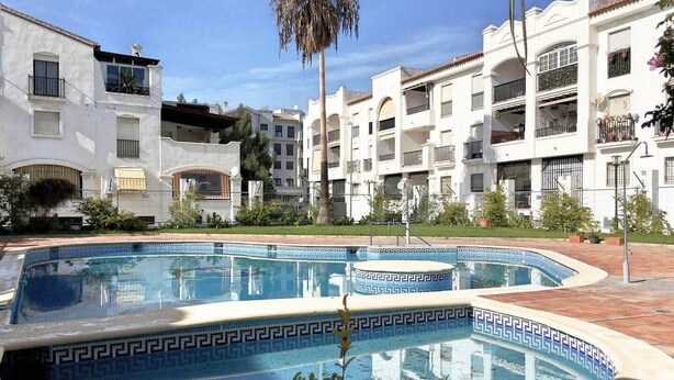 A swimming pool with a geometric tile pattern, surrounded by multi-story white buildings with balconies and windows. - Comeback La Casa Sin Nombre (Motril)