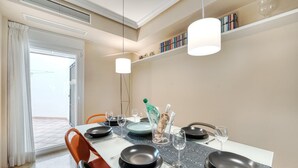 A dining area with a table set for four, including plates, cutlery, and glasses. A bottle of champagne is on the table, and there are books on a shelf above.