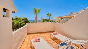 A rooftop terrace with two sun loungers, a small table with a chair, and a view of the sea and palm trees.