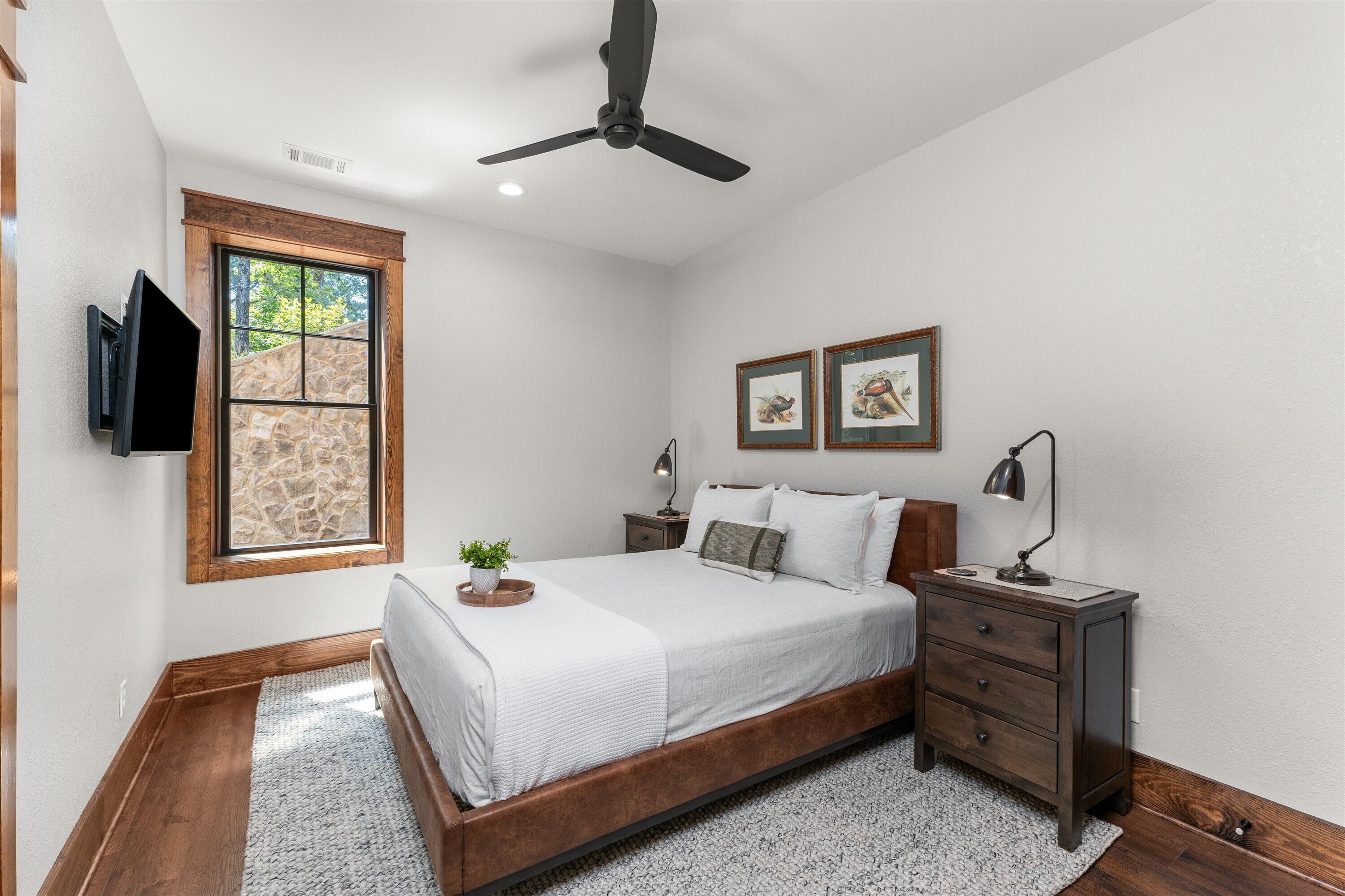 A bedroom with a bed, a nightstand, a TV mounted on the wall, and a window with a stone wall view.