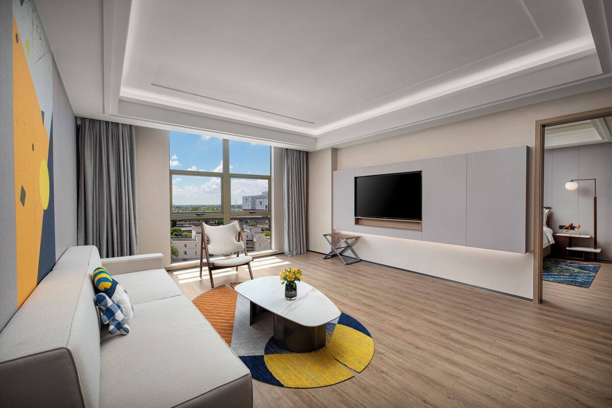A modern hotel room with a large window, a flat-screen TV, a sofa, a coffee table, and a small side table.