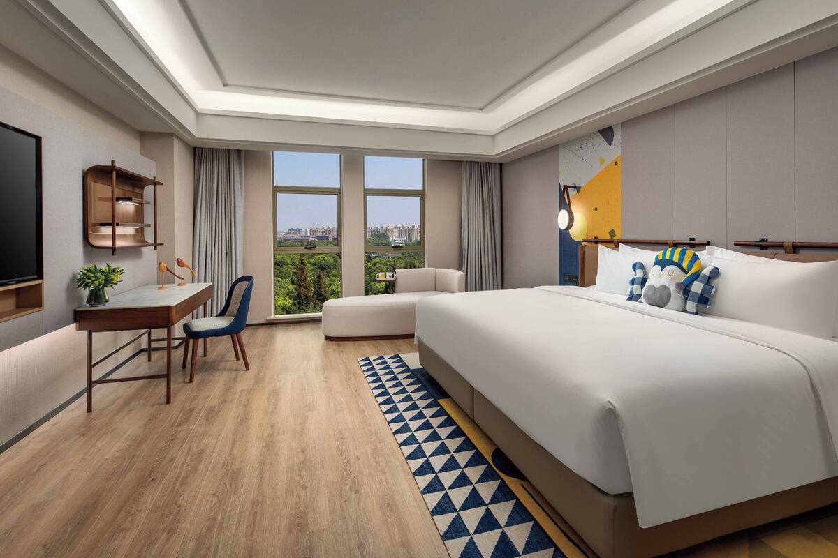 A modern hotel room with a large bed, a desk with a chair, a TV, and a view of the city.