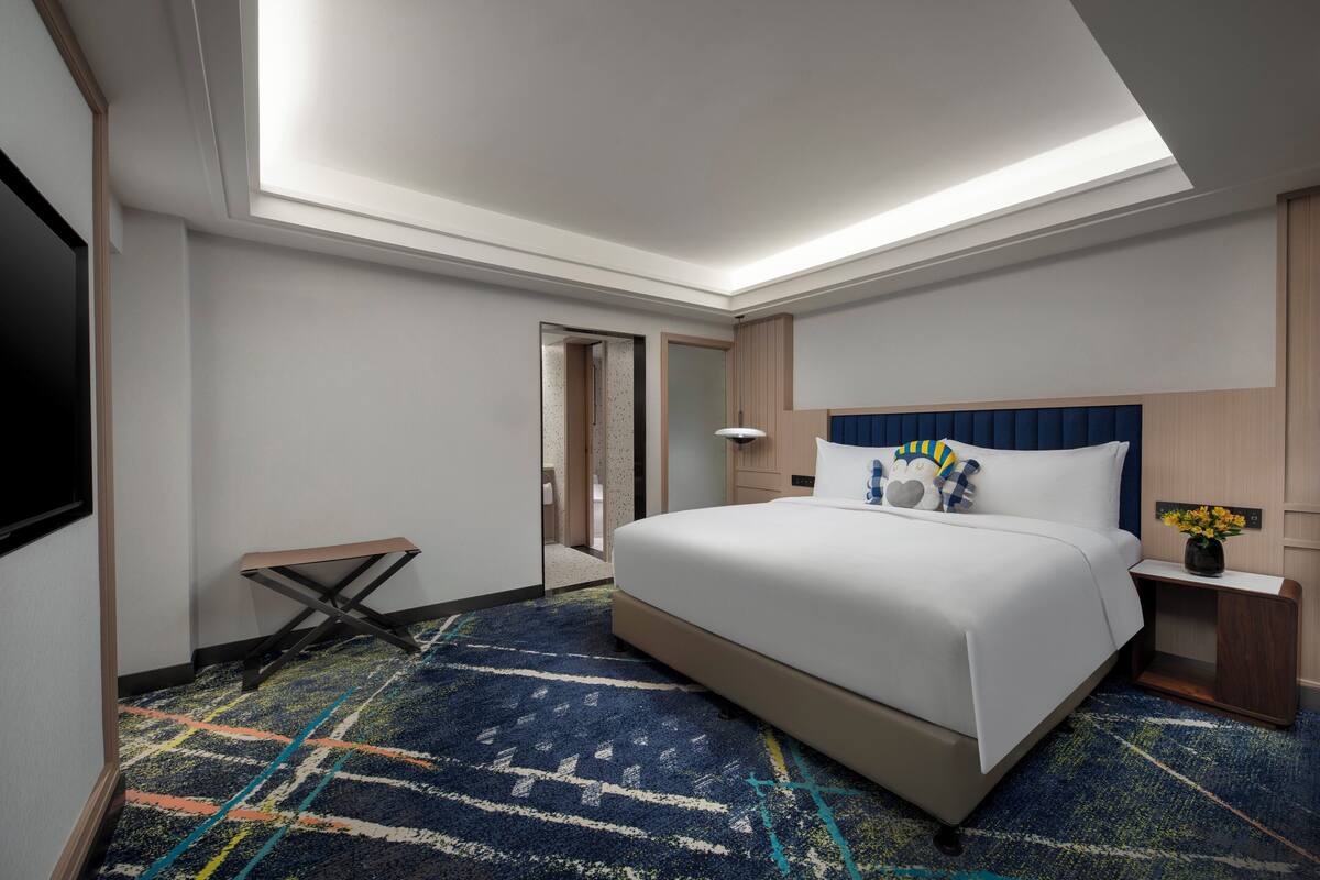 A hotel room with a large bed, a flat-screen TV, a bedside table with a lamp, and a patterned carpet.