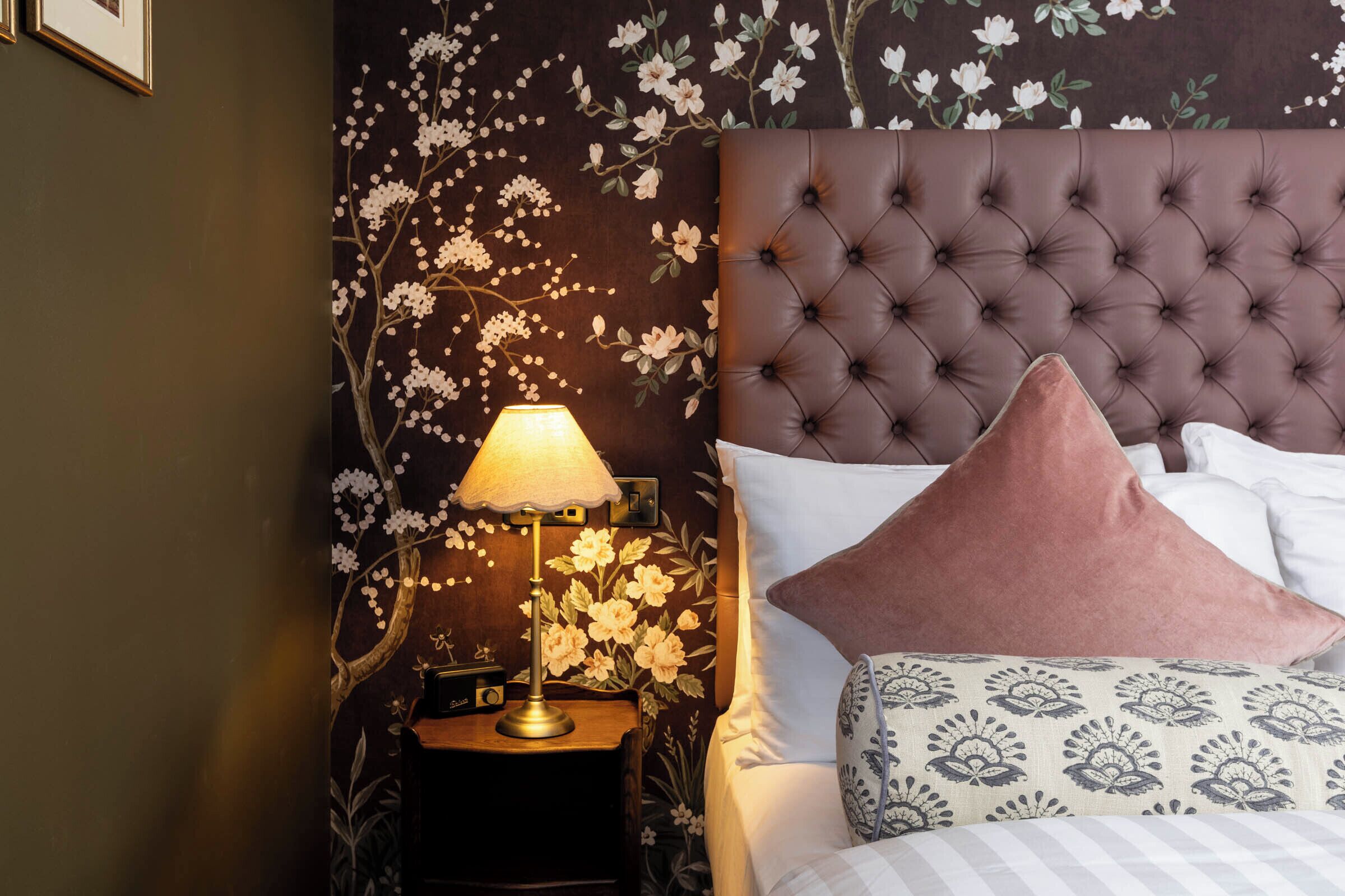 A bedroom with a tufted headboard, a bedside table with a lamp, and floral wallpaper.