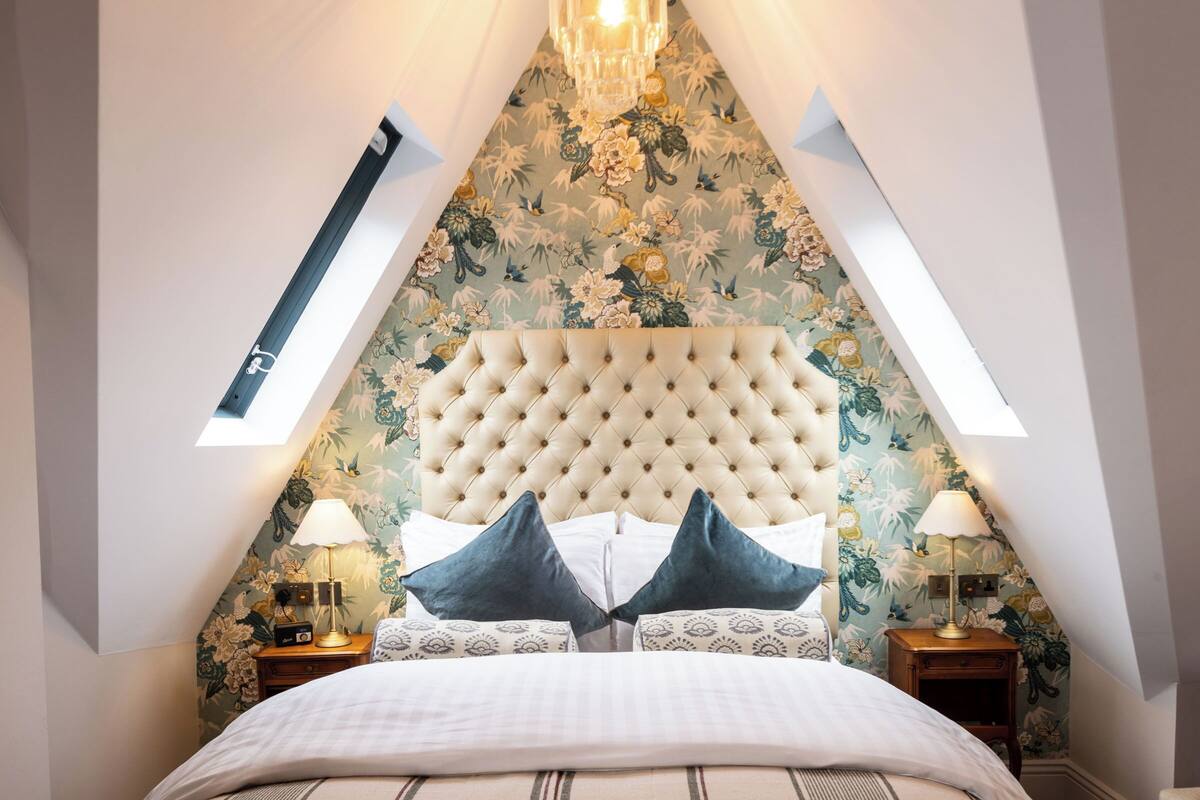 Un dormitorio con cabecera capitonada, tragaluz, papel tapiz floral y cama con ropa de cama estampada.