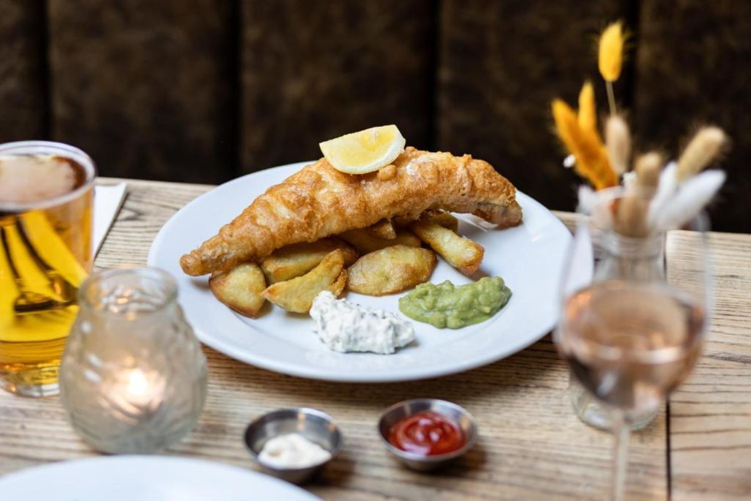 A plate of fish and chips with lemon, tartar sauce, and mushy peas, accompanied by a glass of beer and a small bottle of sauce.