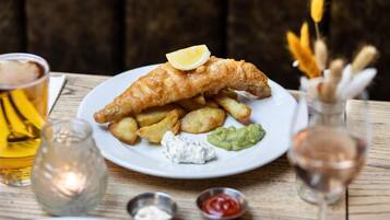 A plate of fish and chips with lemon, tartar sauce, and mushy peas, accompanied by a glass of beer and a small bottle of sauce.