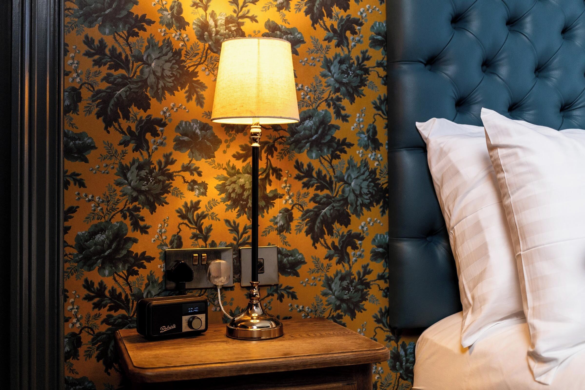 A bedroom with a floral wallpaper, a tufted headboard, a bedside lamp, a radio, and a white pillow.