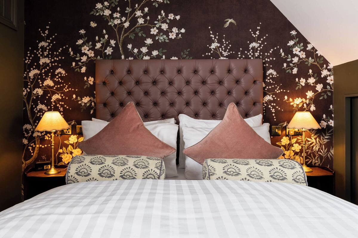 Un dormitorio con una cabecera capitonada, dos mesitas de noche con lámparas, almohadas estampadas y un diseño de papel tapiz floral.