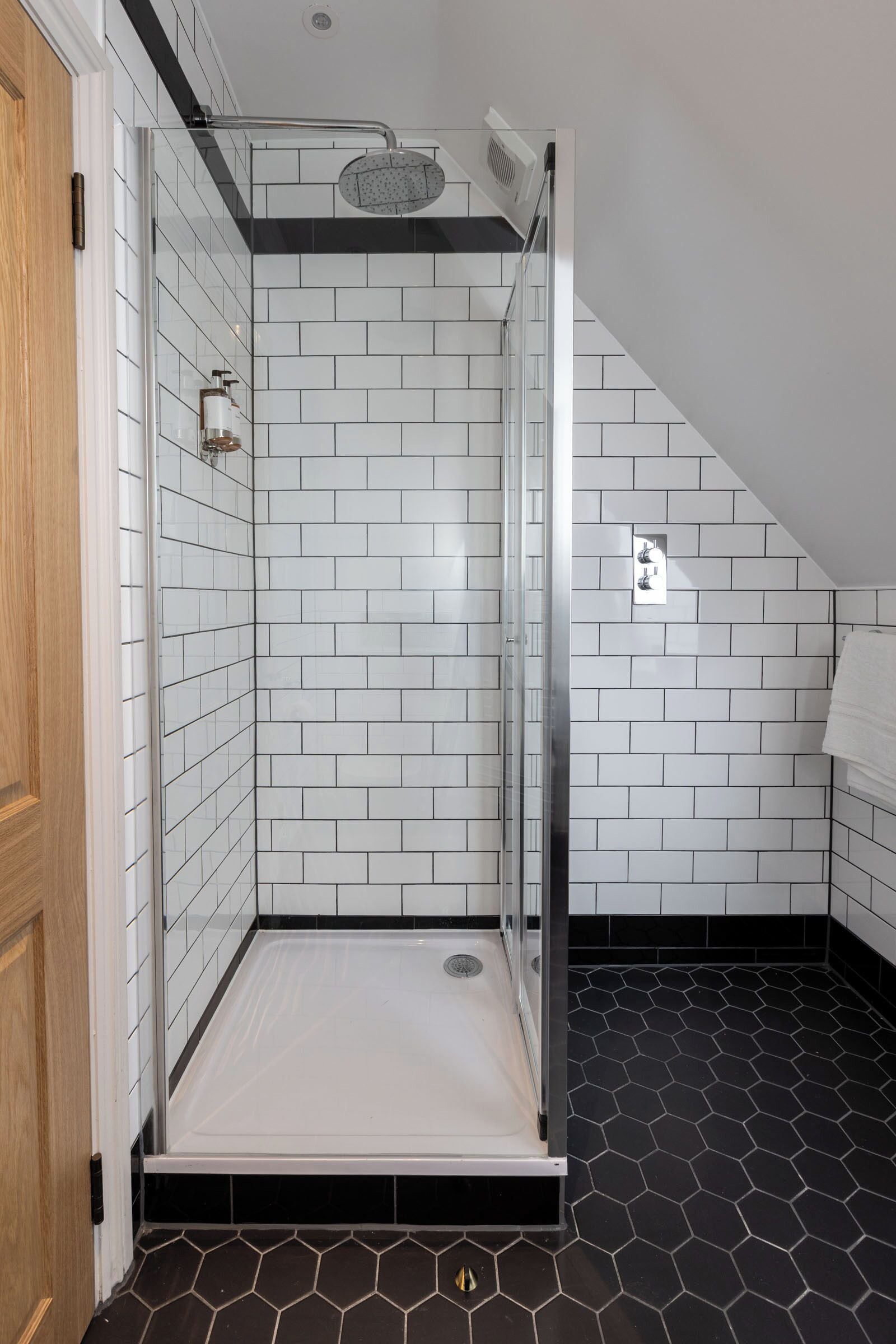 Een douche met witte tegels, een glazen deur en een vloer van zwarte honingraten.