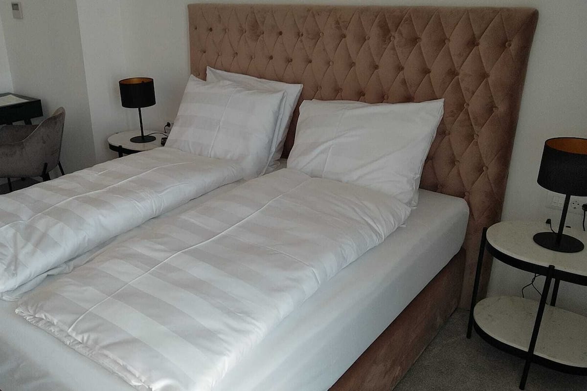 A neatly made twin bed with a tufted headboard, a bedside table, a chair, and a desk with a lamp.