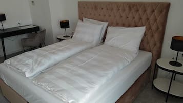 A neatly made twin bed with a tufted headboard, a bedside table, a chair, and a desk with a lamp.
