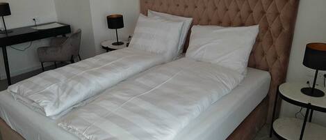 A neatly made twin bed with a tufted headboard, a bedside table, a chair, and a desk with a lamp.
