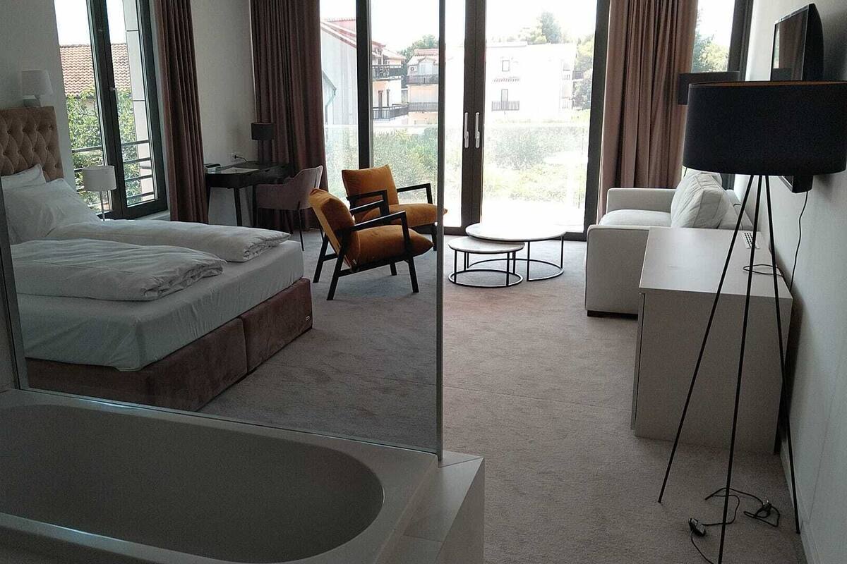 A modern hotel room with a large bathtub, a bed, a desk, a chair, a small table, and a TV.