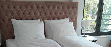 A neatly made bed with a tufted headboard, white bedding, and a view of greenery through a window.