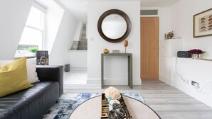 A modern living room with a black leather sofa, a round mirror, a console table with decorative items, and a view of the kitchen through an open door. - Beautiful Notting Hill Apt (Greater London)