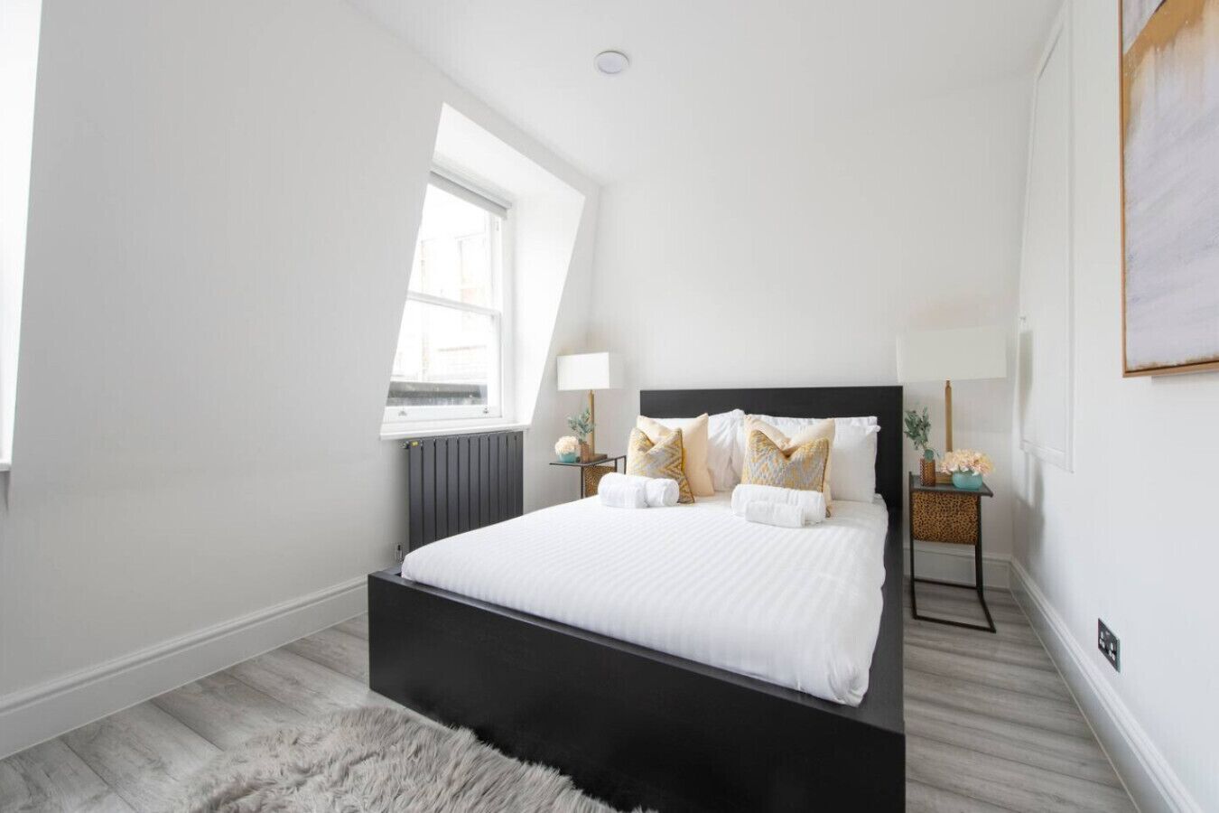 A modern bedroom with a black bed frame, white bedding, and decorative pillows. There is a window providing natural light and a small bedside table with a lamp and flowers.