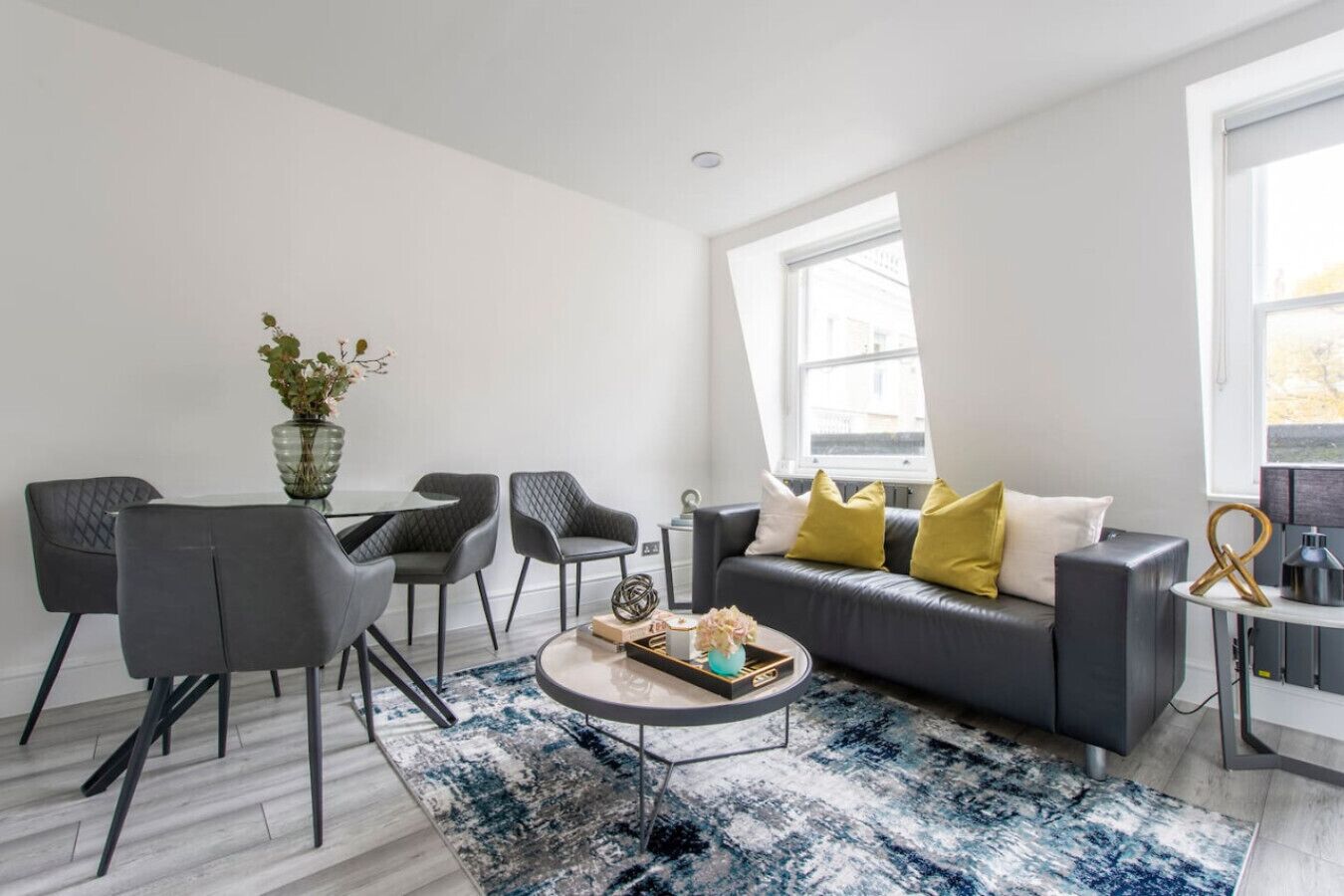 Een moderne woonkamer met een grijze bank, zwarte stoelen, een salontafel en een blauw gestreept vloerkleed.