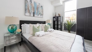 A modern bedroom with a large bed, a nightstand with a lamp, a bookshelf, and a window with a view of the outside. - Beautiful Notting Hill Apt (Greater London)