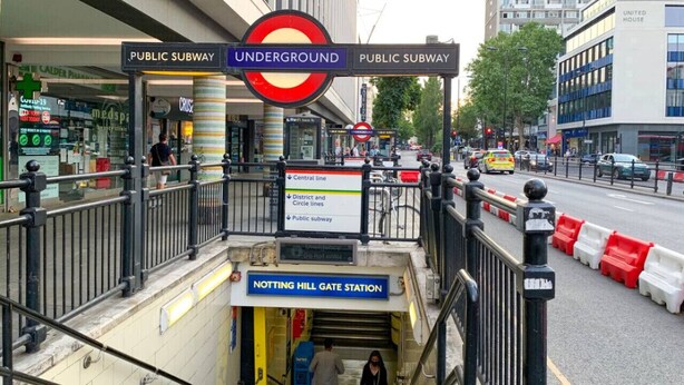 A subway entrance with a sign for Notting Hill Gate Station, surrounded by a street with cars and buildings.