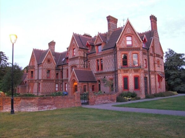 Stately building with a mix of brickwork and decorative elements, set amidst a well-manicured lawn and paved pathway.