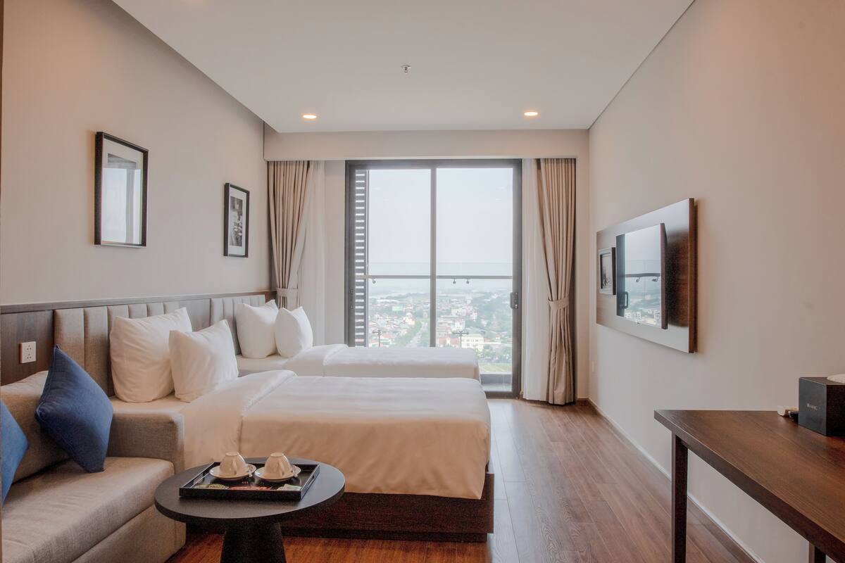 Habitación de hotel con dos camas, un sofá, una mesita con bandeja, televisión y un ventanal con vistas a la ciudad.