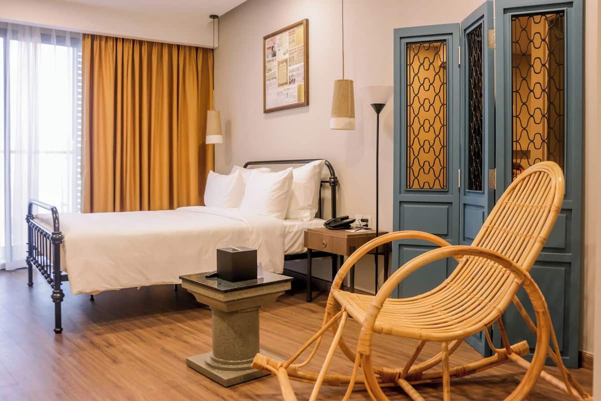 Una habitación de hotel con una cama, una mesita de noche, una silla de mimbre y una puerta con un panel decorado.