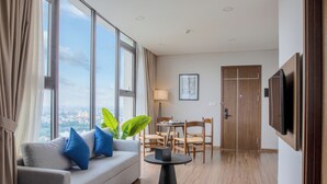 A modern hotel room with a sofa, a dining table, chairs, a TV, and large windows offering a city view. - Mandala Hotel & Suites Hai Duong (Hai Phong)
