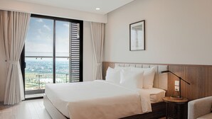 A hotel room with a large bed, a bedside table with a lamp, a framed picture on the wall, and a view of the city through the window. - Mandala Hotel & Suites Hai Duong (Hai Phong)