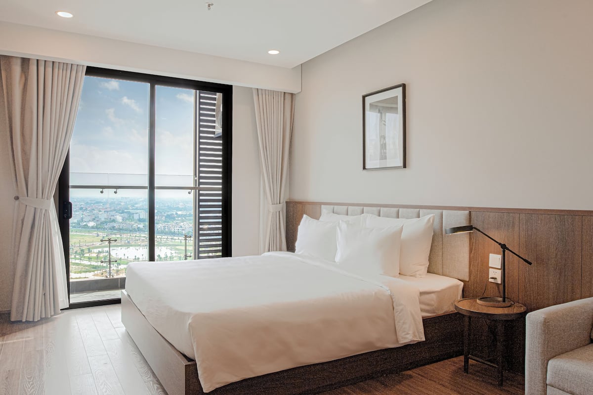 Habitación de hotel con una cama grande, una mesita de noche con lámpara, un cuadro enmarcado en la pared y vistas de la ciudad a través del ventanal.