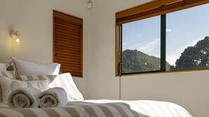 A bedroom with a bed, pillows, a window with a view of a forested hill, and a wall-mounted light fixture. - Heart of Mt Maunganui (Mount Maunganui)