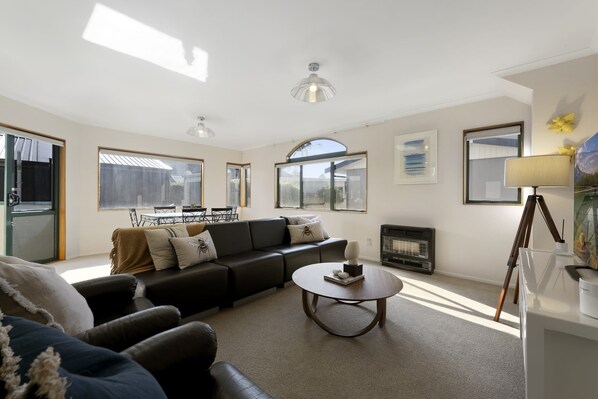 A living room with a black sofa, a round coffee table, and a fireplace. There are large windows and a door leading outside.