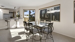 A modern kitchen with white cabinets, a microwave, and a coffee maker. There is a dining area with a glass table and metal chairs. - Heart of Mt Maunganui (Mount Maunganui)