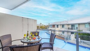 A balcony with wicker chairs, a glass table with food and drinks, and a pool view.
