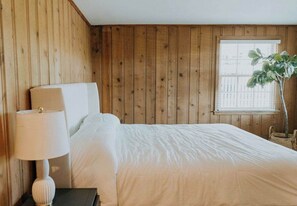 A bedroom with wooden paneling, a bed with white bedding, a bedside lamp, and a window with a plant. - Palmetto Cottage (Wrightsville Beach)