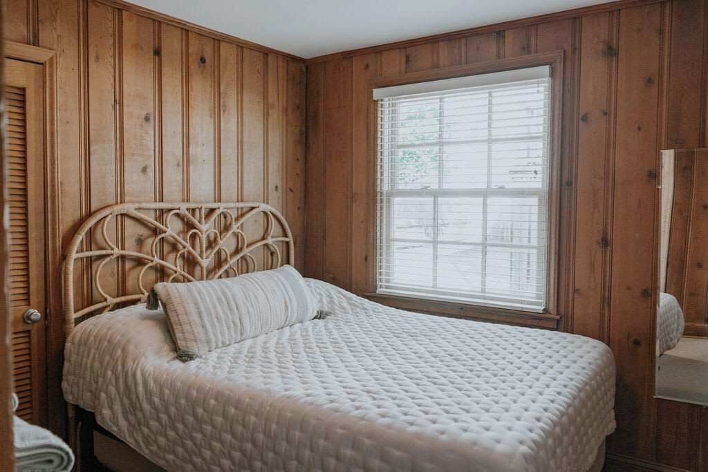 Een kamer met houten lambrisering, een bed, een raam met luiken en een deur.