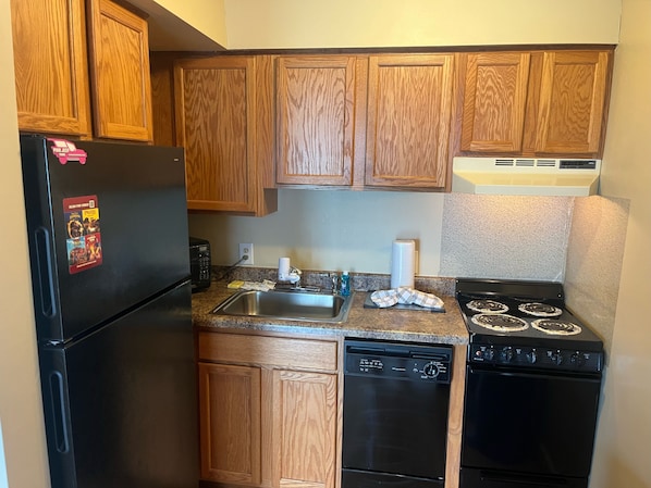 A compact kitchen with wooden cabinets, a black refrigerator, a dishwasher, a stove, and a microwave.