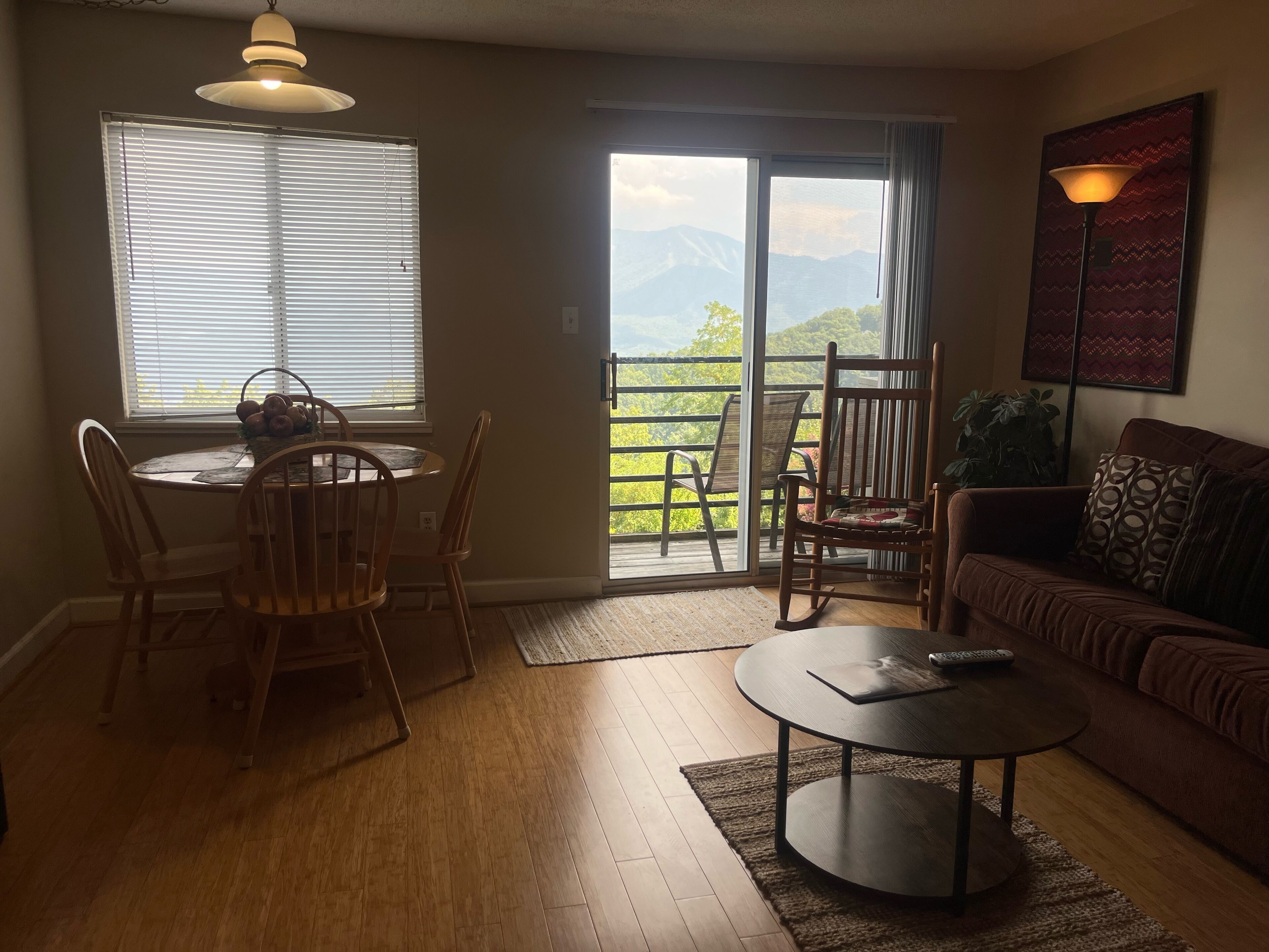 A living room with a dining table, chairs, a sofa, a coffee table, and a view of mountains through a sliding door.