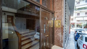 Entrada de hotel com porta de vidro, degraus de mármore e área de estacionamento visível.