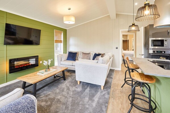 A modern living room with a sofa, a coffee table, and a TV mounted on a green wall. There is a kitchen area with a microwave and a stove. - Host Stay Eller Beck - The Cabin in the Woods (Whitby)