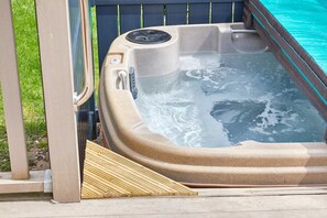 A hot tub with a control panel, a wooden step leading into it, and a pool in the background.