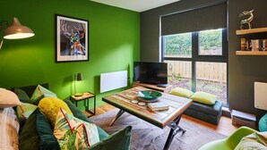 A modern living room with green walls, a large window, a flat-screen TV, a wooden coffee table, and a bookshelf. - Host Stay Forest Green Lodge (Alnwick)